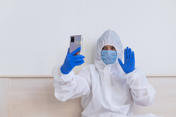 Een vrouwelijke arts in een beschermend pak maakt een videogesprek op een smartphone - Foto, afbeelding