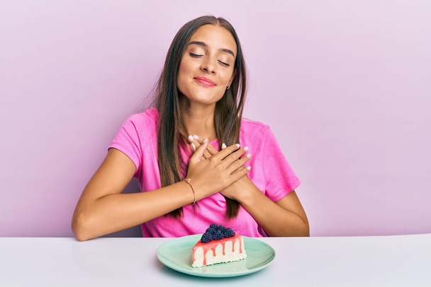 Giovane donna ispanica che mangia cheesecake seduta sul tavolo sorridente con le mani sul petto, gli occhi chiusi con gesto grato sul viso. concetto di salute.  - Foto, immagini