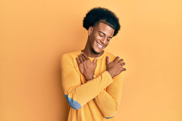 Afro-Amerikaanse man met afrohaar in casual kleding die zichzelf omhelst gelukkig en positief, glimlachend vol zelfvertrouwen. zelfliefde en zelfzorg  - Foto, afbeelding