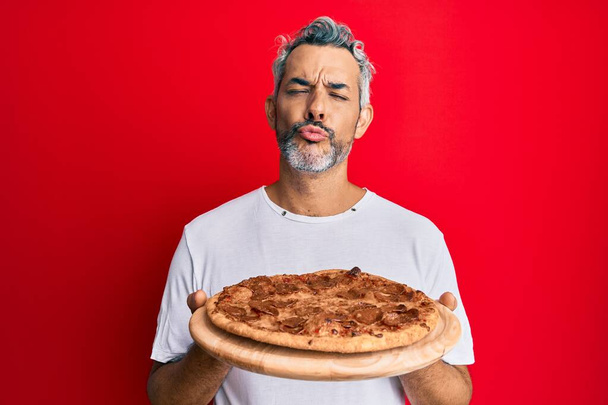 Grauhaariger Mann mittleren Alters, der eine italienische Pizza in die Kamera hält und einen Kuss bläst, der schön und sexy ist. Liebesausdruck.  - Foto, Bild