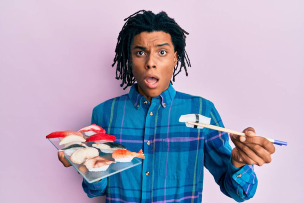 Young african american man eating sushi using chopsticks in shock face, looking skeptical and sarcastic, surprised with open mouth  - Photo, Image