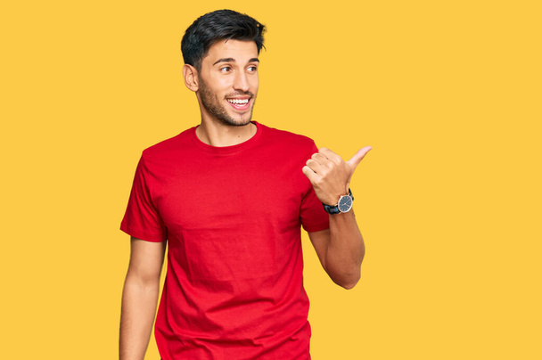 Young handsome man wearing casual red tshirt smiling with happy face looking and pointing to the side with thumb up.  - Photo, Image