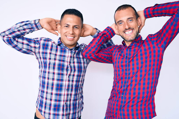 Jeune couple gay de deux hommes portant des vêtements décontractés relaxant et étirant, les bras et les mains derrière la tête et le cou souriant heureux  - Photo, image