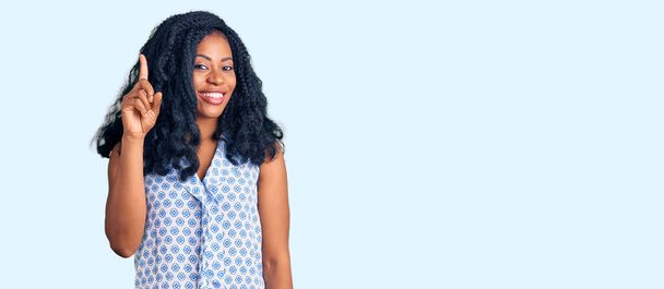 Piękna Afroamerykanka ubrana w luźną letnią koszulę, pokazująca i wskazująca palcem numer jeden, uśmiechnięta pewna siebie i szczęśliwa..  - Zdjęcie, obraz