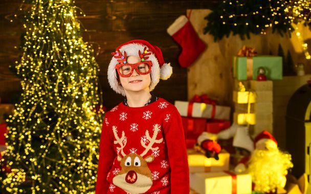 Kid wear santa hat and christmas sweater. Santa brought me gifts. December tradition. Sale discount. Real happiness. Child happy excited near christmas tree. Merry christmas. Happy childhood concept - Φωτογραφία, εικόνα