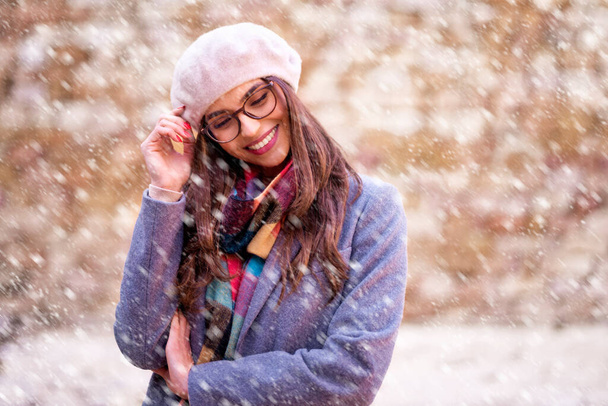 Portrait shot of attractive young woman wearing beret hat and winter coat while standing outside and enjoy falling snow. - Foto, Imagen