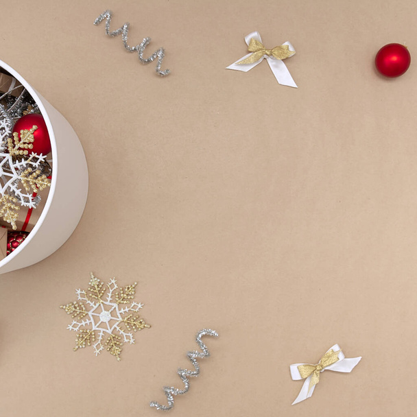Eine weiße runde Schachtel mit Weihnachtsspielzeug, daneben Dekorationen aus rot, weiß, gold und silber auf beigem Hintergrund - Foto, Bild