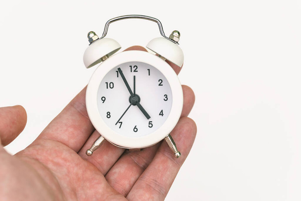 Der weiße Wecker liegt in der Handfläche einer Person. Die Stundenzeiger zeigen fünf Uhr morgens oder abends an. Ende des Arbeitstages. Der Begriff der Arbeit. Zeitkonzept. - Foto, Bild