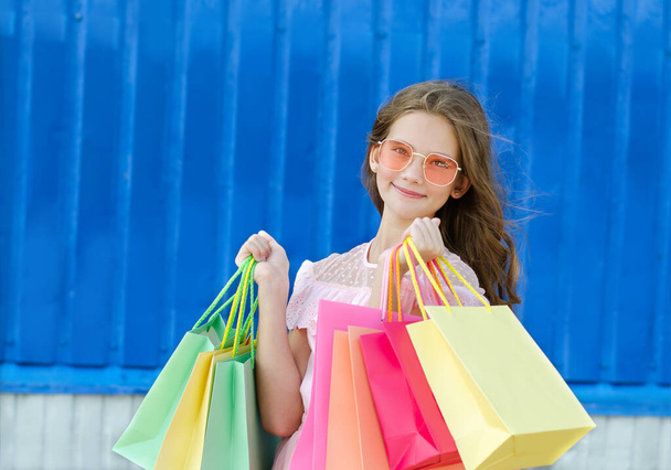 Beautiful happy smiling little girl child in sunglasses is holding shopping bags near shopping mall outdoors. Lifestyle concept - Zdjęcie, obraz