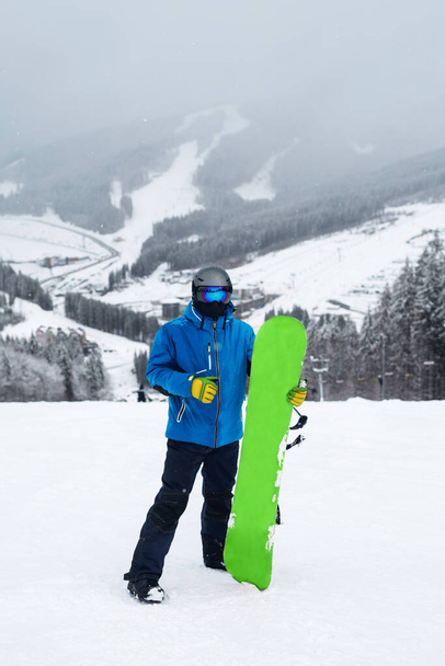 Парень держит зеленый сноуборд рядом с ногой зимой. Сноубордист в синей куртке на фоне снежных гор - Фото, изображение