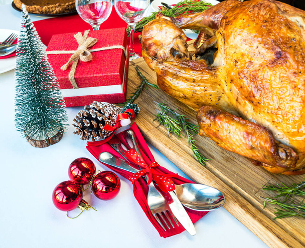 Whole roast turkey and rosemary on a wooden cutting board with festive decoration and cutlery setting around it and pumpkin pie on the back on white tablecloth and red table runner - Photo, Image