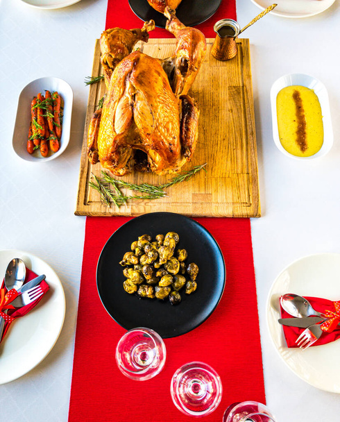 Christmas dinner table concept of whole roast turkey on wooden cutting board with brussel sprout, glazed carrots, mashed potatoes, gravy, tableware on white tablecloth and red table runner - Photo, Image