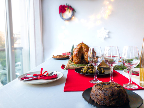 Christmas dinner table concept of christmas pudding, wine and glasses, cutlery setting, brussel sprout, roast turkey, pumpkin pie with festive christmas decoration on white tablecloth,red table runner - Photo, Image