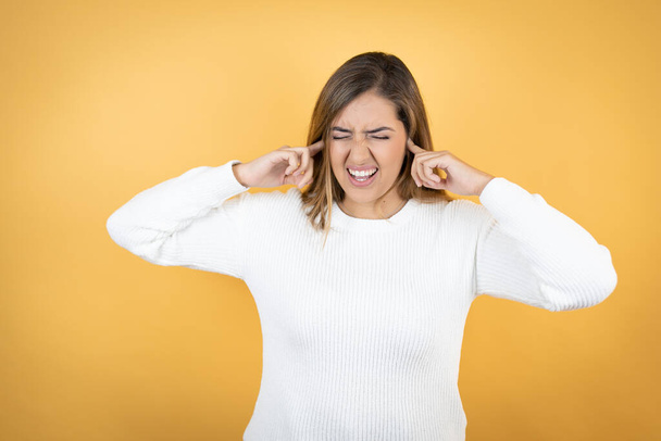 Young caucasian woman over isolated yellow background covering ears with fingers with annoyed expression for the noise of loud music. Deaf concept. - Photo, Image