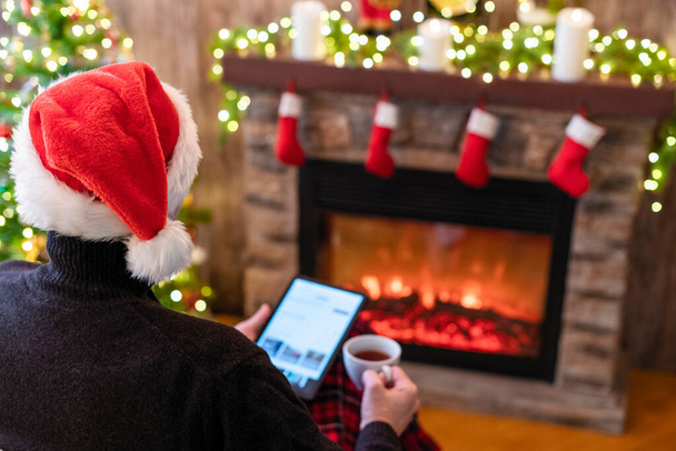 Christmas. Man in santa claus hat with tablet and cup of tea sitting near christmas tree and fireplace. Concept of online service in holidays. Back view. - Foto, Imagem
