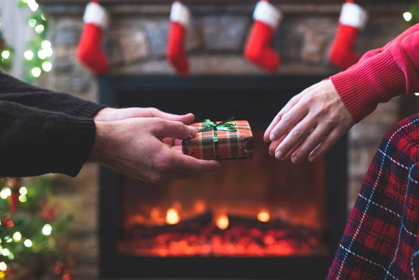 Man giving Christmas and New Year Gift box to woman in front of christmas tree, fireplace with flame. Xmas concept. - 写真・画像