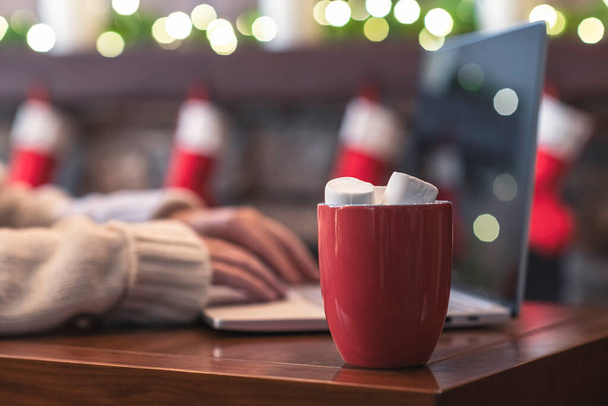 Lonely woman freelancer enjoying with cup of hot cocoa and marshmallow working on laptop near christmas fireplace with decoration of light bulbs. - Φωτογραφία, εικόνα
