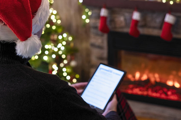 Christmas. Man in santa claus hat with tablet sitting near christmas tree and fireplace. Concept of online service in holidays. Back view. - 写真・画像