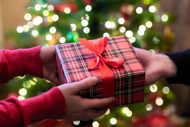 Mann schenkt Frau gegenüber Weihnachtsbaum Weihnachts- und Neujahrsgeschenkbox mit glühenden Glühbirnen. Weihnachtskonzept. Aus nächster Nähe. Selektiver Fokus - Foto, Bild