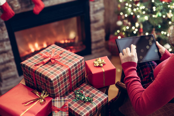 Christmas. Woman in sweater using tablet for searching gift ideas sitting at table near fireplace and christmas tree. Concept - Valokuva, kuva