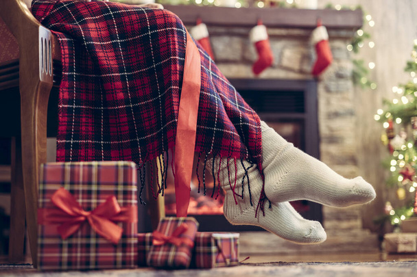 Las piernas de la mujer en un invierno calcetines cubiertos a cuadros sentarse y relajarse en el sillón cerca de la chimenea y el árbol de Navidad pakking cajas de regalo para la familia. Vista inferior. - Foto, imagen