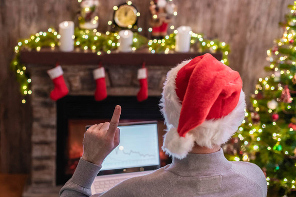 Man freelancer in santa claus hat working on laptop with graphs and charts raising a finger up sitting near christmas tree and fireplace. - Fotografie, Obrázek