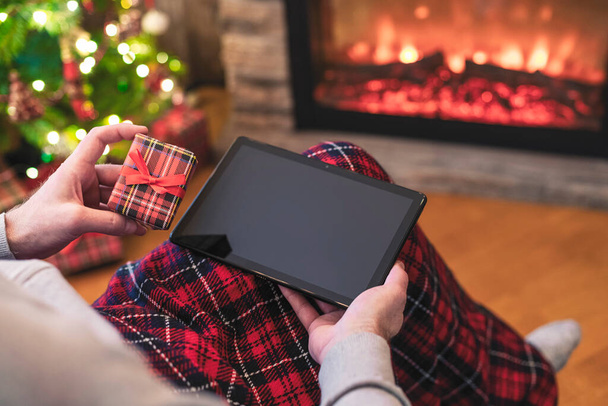 Christmas. Man using tablet for searching gift ideas sitting with ready gift boxe near fireplace and christmas tree. Concept - Foto, Imagen