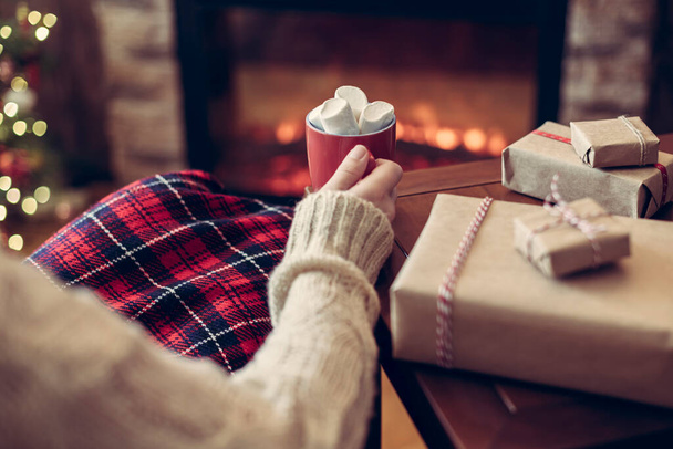 Woman covered plaid sitting and relaxation near fareplace and christmas tree with cup of cocoa and marshmallows after finishing packing gift boxes for family.  - 写真・画像