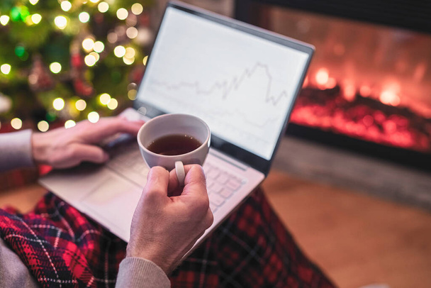 Man freelancer working on laptop with graphs and charts on screen sitting near christmas tree and fireplace with cup of tea. - Valokuva, kuva