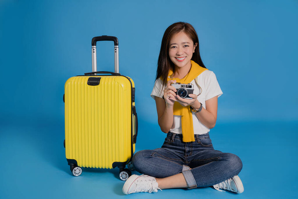 Молода азіатка - мандрівниця сидить біля жовтої валізи. Вона тримає фотоапарат на синьому фоні. - Фото, зображення