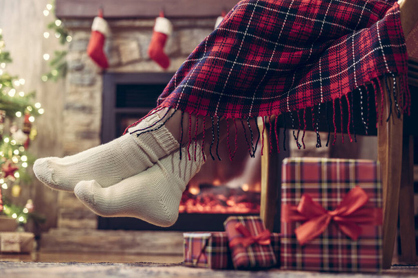 Woman legs in a winter socks covered plaid sitting and relaxation on armchair near fireplace and christmas tree pakking gift boxes for family. Bottom view. - Фото, изображение