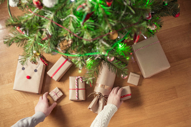 Vrouw en man die met de hand geschenken inpakken in een kraftpapier onder de kerstboom. Cadeaus voor familie. Bovenaanzicht. Vlakke plaat. - Foto, afbeelding