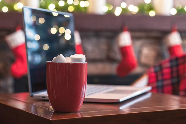 Laptop, cup of hot chocolate with marshmallow on the table at christmas fireplace with decoration of light bulbs. Close up. - Φωτογραφία, εικόνα