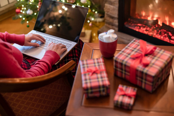 Navidad. Mujer que utiliza el ordenador portátil para la búsqueda de ideas de regalo sentado en la mesa con cajas de regalo listo y taza de cacao y malvaviscos cerca de la chimenea y el árbol de Navidad. Concepto - Foto, Imagen