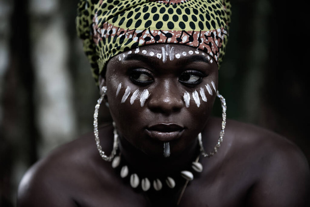 African woman sitting in the Aburi forest in Ghana with white painting on her face and headdress - Photo, Image