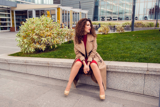 Brunette young woman in beige coat and high heel shoes sitting outdoors - Photo, Image