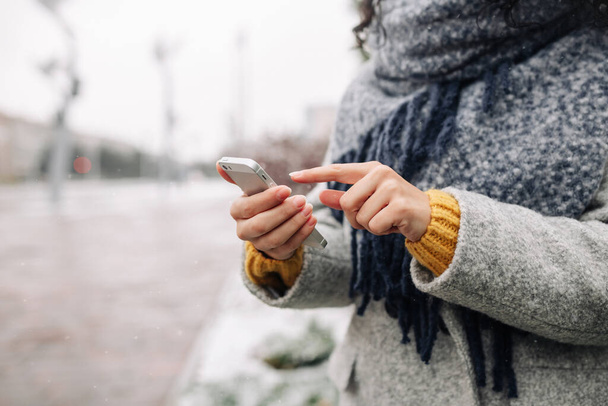 Νεαρή γυναίκα χρησιμοποιεί το κινητό της τηλέφωνο σε ένα χιονισμένο χειμερινό πάρκο. Κλείσιμο του γυναικείου ελέγχου ειδήσεων και γραπτών μηνυμάτων στο κινητό της κατά τη διάρκεια της κρύας χειμερινής περιόδου. Έννοια για τα γκάτζετ των ανθρώπων - Φωτογραφία, εικόνα