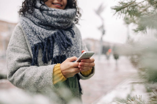 Junge Frau mit ihrem Handy in einem verschneiten Winterpark. Nahaufnahme von Frauen, die während der kalten Wintersaison Nachrichten checken und SMS auf ihrem Handy schreiben. Volkstümliches Spielzeugkonzept - Foto, Bild