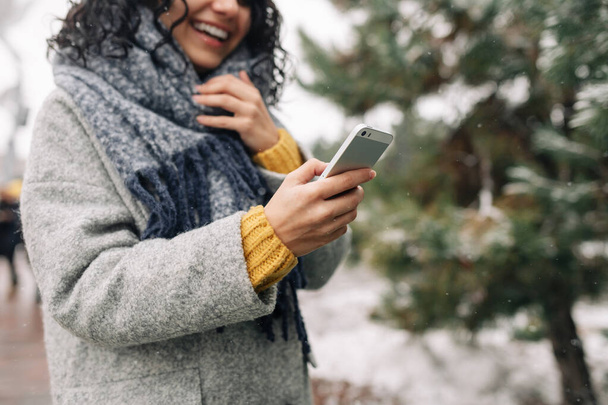 Young woman using her mobile phone at a snowy winter park. Closeup of female checking news and texting on her cellphone outdoor during cold winter season. Peoples' gadgets concept - Foto, Imagem