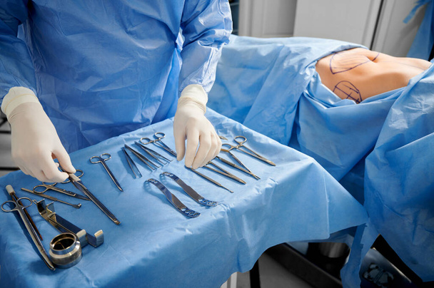滅菌手袋の外科医の閉鎖準備医療機器を取得します。医師がツールを準備しながら、皮膚にマークを持つ女性患者は、医療ベッドの上に横たわっています.整形手術の準備の概念. - 写真・画像