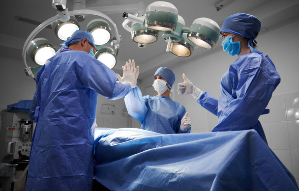 Artsen staan klaar voor de patiënt na een succesvolle operatie. Plastisch chirurg geeft high five aan assistent. Tweede assistent doet duimen omhoog gebaar. Concept van geneeskunde en succesvol teamwerk. - Foto, afbeelding