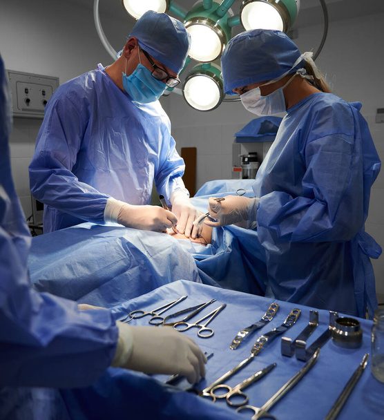 Медицинская команда в хирургических костюмах проводит эстетическую операцию в операционной. Врачи в стерильных перчатках и защитных масках делают косметическую операцию. Концепция пластической хирургии. - Фото, изображение