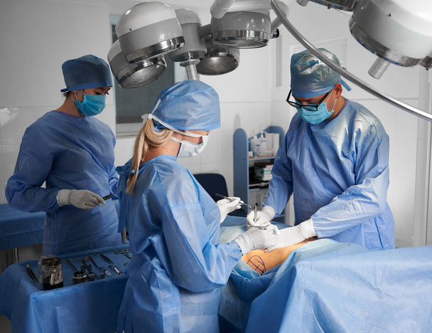 Orvosok csoportja kék sebészeti egyenruhában, akik kozmetikai műtétet végeznek a műtőben. A sebész csapat védőmaszkot, steril kesztyűt és orvosi sapkát visel. A plasztikai sebészet fogalma. - Fotó, kép
