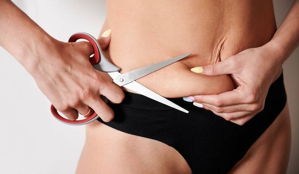 Close up of woman hands cutting belly fat with scissors. Female in black underwear trying to get rid of excess weight. Concept of weight loss, diet and overweight. - Фото, зображення