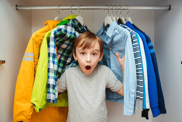 Překvapený roztomilý chlapec hledá oblečení ve skříni. Preteen chlapec si vybírá oblečení v šatníku doma. Dítě schované mezi šaty ve skříni. - Fotografie, Obrázek