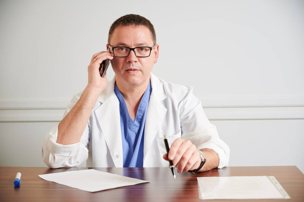 Un cirujano serio con gafas conversando por teléfono, sentado en la mesa con papeles. Médico varón con bata de laboratorio blanca sosteniendo pluma. Concepto de personal médico, de comunicación y sanitario. - Foto, imagen
