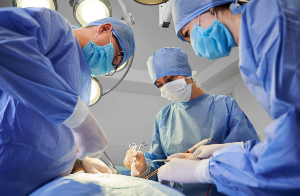 Sebész csapat kék orvosi öltönyben, orvosi műszerekkel és plasztikai műtéttel. Férfi sebész és női asszisztensek védőmaszkot, steril kesztyűt és sapkát viselnek a modern klinikán - Fotó, kép
