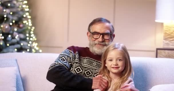 Close up portret wesołej rodziny kaukaskiej starego dziadka z cute little girl siedzi w urządzonym pokoju w pobliżu choinki uśmiecha się i przytula w Sylwestra. Zimowe wakacje. Koncepcja świąteczna - Materiał filmowy, wideo