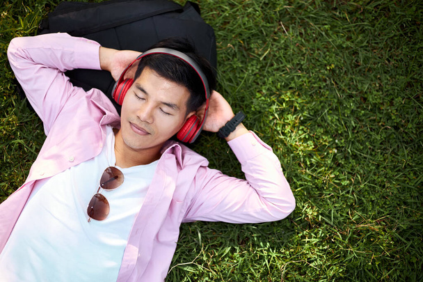 Asijský muž lež poslech hudby se sluchátky na trávě relaxační. - Fotografie, Obrázek