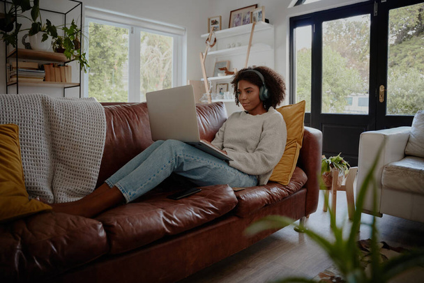 African young woman relaxing on couch while watching movie on laptop with wireless bluetooth headphones - Valokuva, kuva